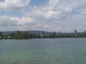 Na Prírodnom kúpalisku Vajnorské jazerá, nádhernej bratislavskej prírodnej vodnej oáze, pláva skupinka ľudí.