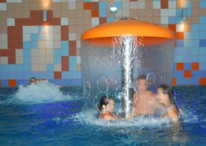 Skupina ľudí hrajúcich sa v bazéne s vodnou fontánou na Krytá plaváreň hotela Nivy v Bratislave.